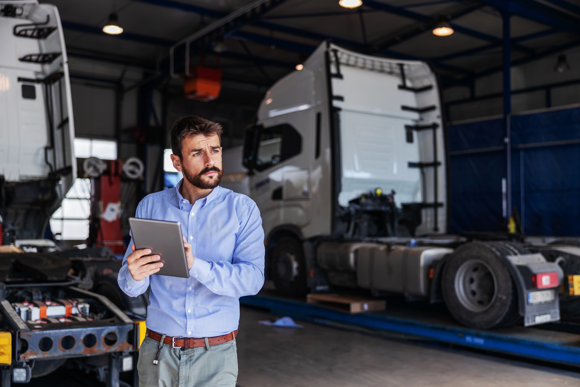 Young serious bearded CEO standing in garage of shipping firm and using tablet. In background are trucks.